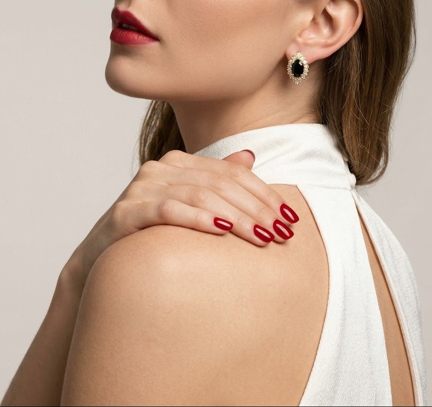 A close-up lifestyle shot of a woman wearing a small black and white teardrop-shaped stud earring, with her hand resting on her shoulder. She is wearing a white sleeveless top and has red nail polish and red lipstick, set against a plain light gray background.