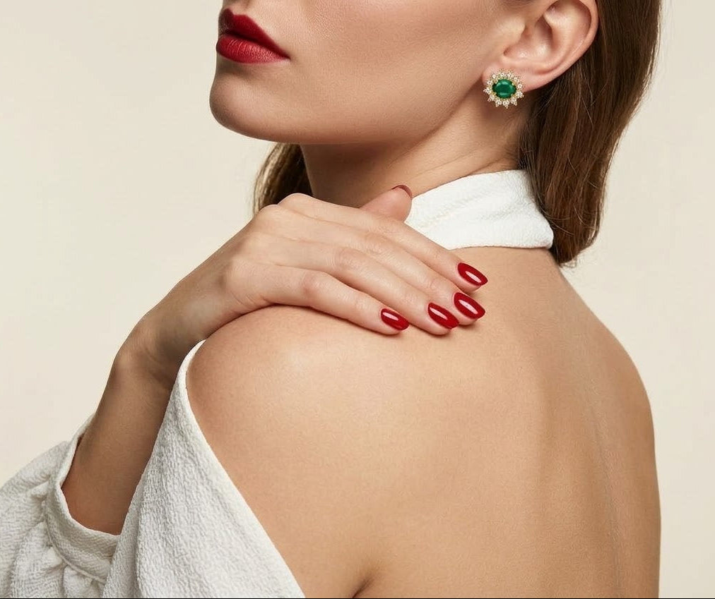A close-up lifestyle shot of a woman wearing a green and gold cluster stud earring, with her hand resting on her shoulder. She has red nail polish and is wearing a white textured top against a warm beige background.