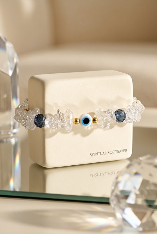 A beaded bracelet with clear faceted crystal-like beads, dark blue round beads, and a central blue-and-white evil eye charm with gold accents, displayed on a cream-colored box labeled 'SPIRITUAL SOOTSAVER'.