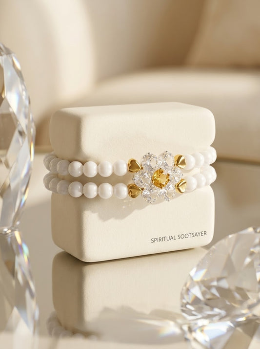 A double-strand white pearl bracelet with a gold and clear crystal flower centerpiece, displayed on a cream-colored jewelry box labeled 'SPIRITUAL SOOTSAVER'.