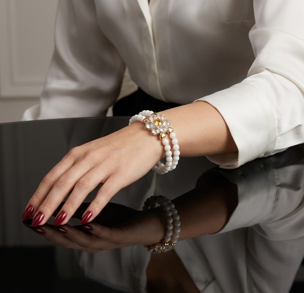 A close-up of a woman's hand and wrist wearing a double-strand pearl bracelet with a gold-toned floral clasp, paired with a white blouse and red manicured nails.