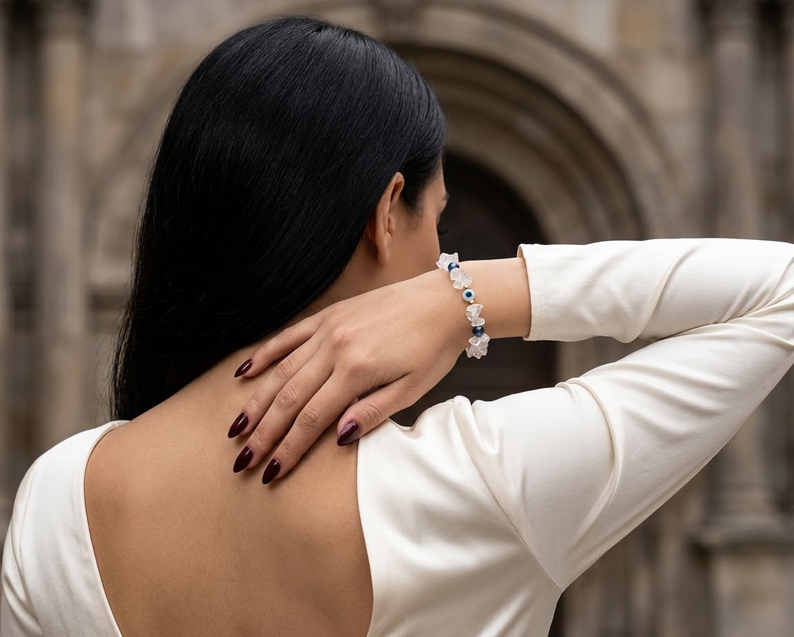 A woman with long black hair, seen from behind, wearing a white long-sleeve top and a beaded bracelet on her wrist, posing with her hand on her neck against a blurred stone archway background.