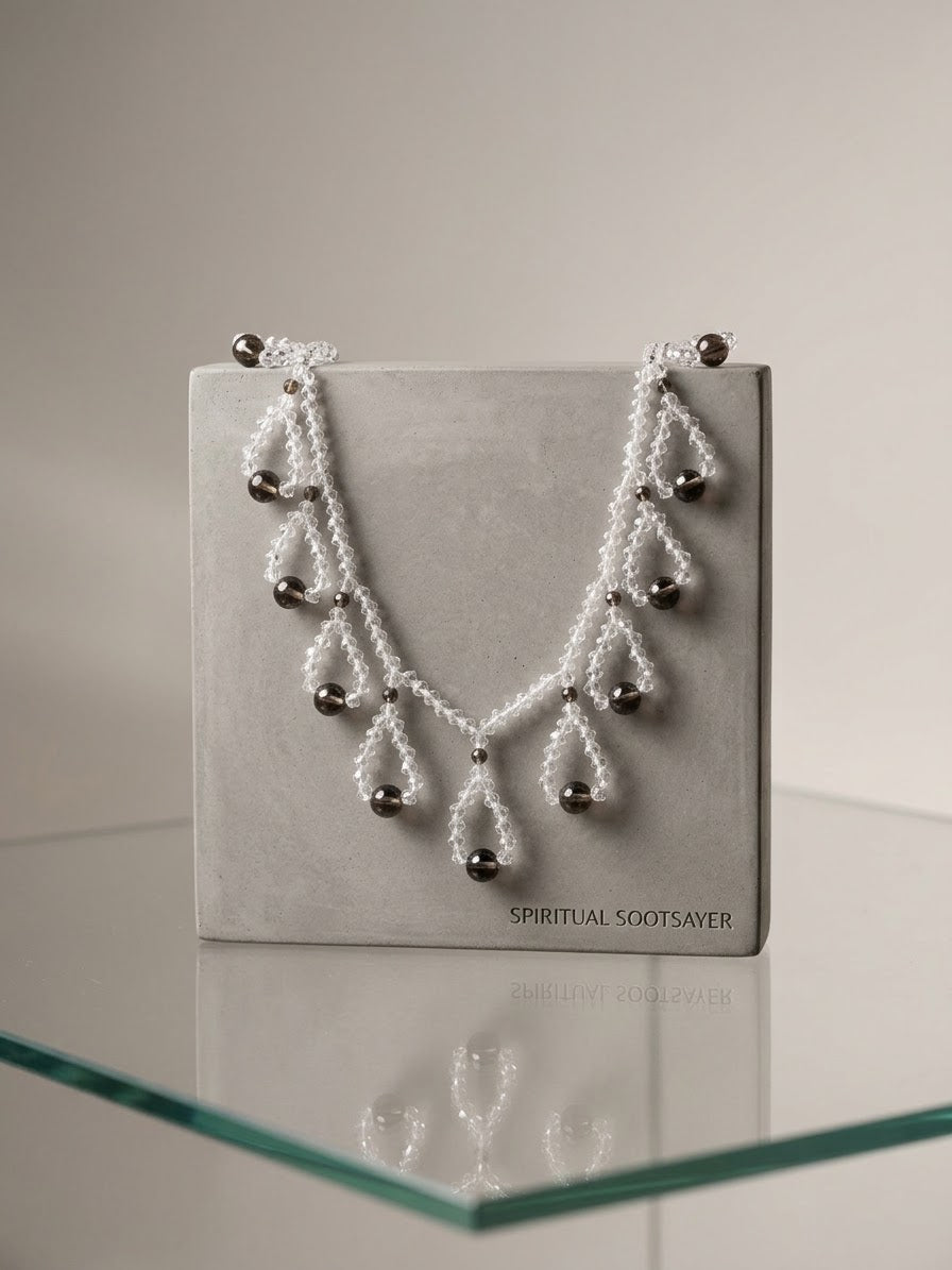 A white beaded necklace with silver-toned metal beads and drop pendants, displayed on a gray jewelry box labeled 'SPIRITUAL SOOTSAVER' on a glass shelf.