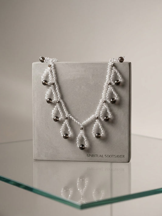 A white beaded necklace with silver-toned metal beads and drop pendants, displayed on a gray jewelry box labeled 'SPIRITUAL SOOTSAVER' on a glass shelf.