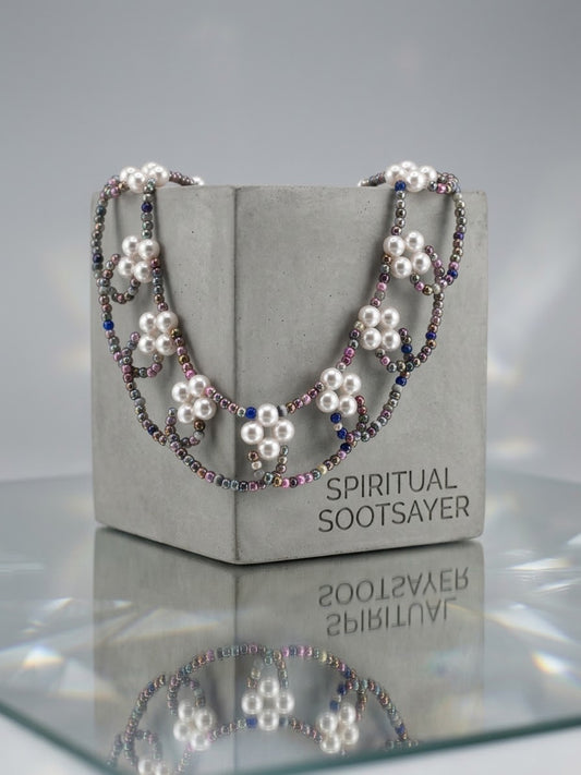 A beaded necklace with white faux pearls and silver-toned beads, draped over a gray display block labeled 'SPIRITUAL SOOTSAYER'.