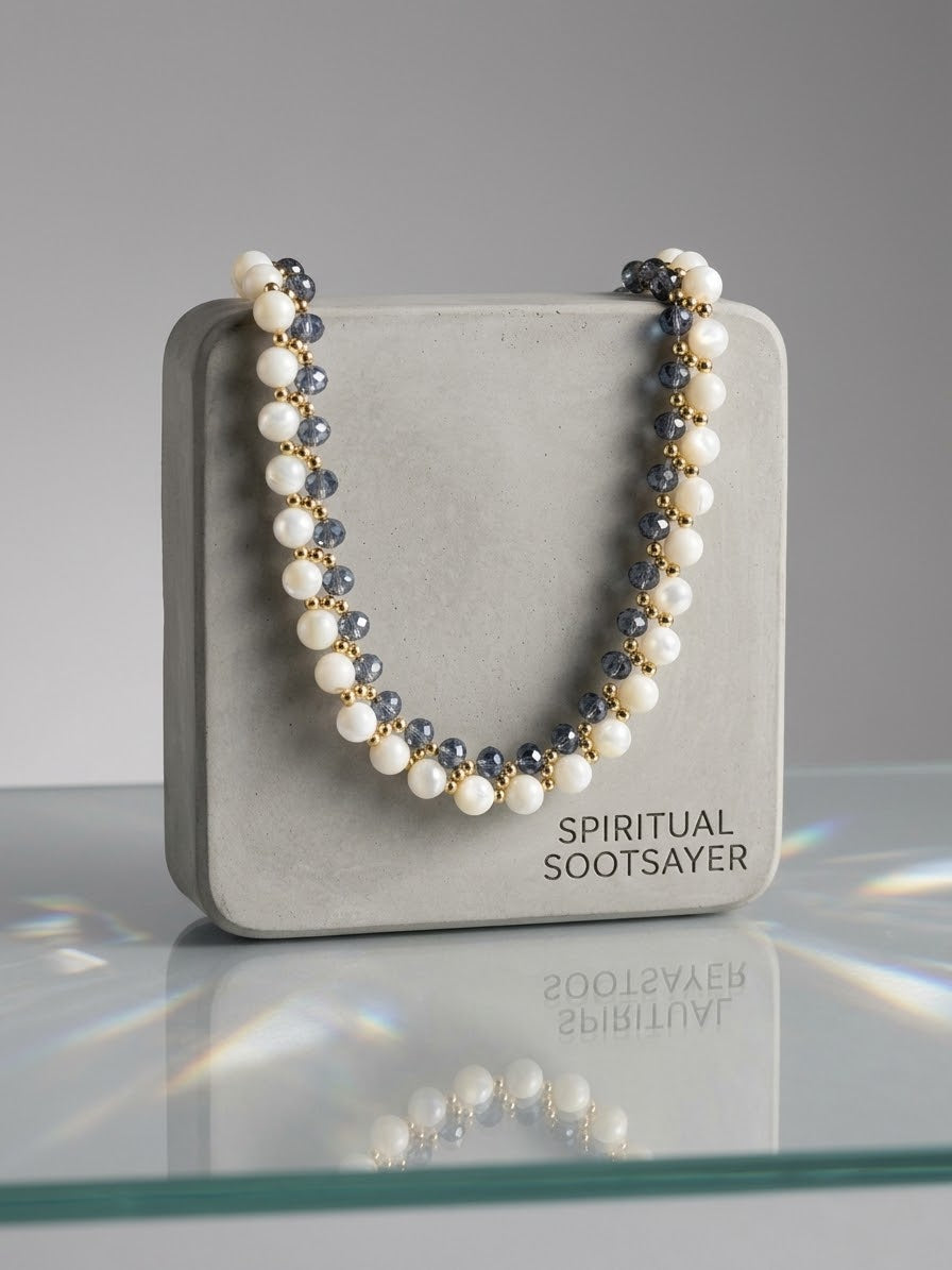 A beaded necklace draped over a gray display box labeled 'SPIRITUAL SOOTSAYER'. The necklace features white pearl-like beads, dark gray faceted beads, and small gold spacer beads.