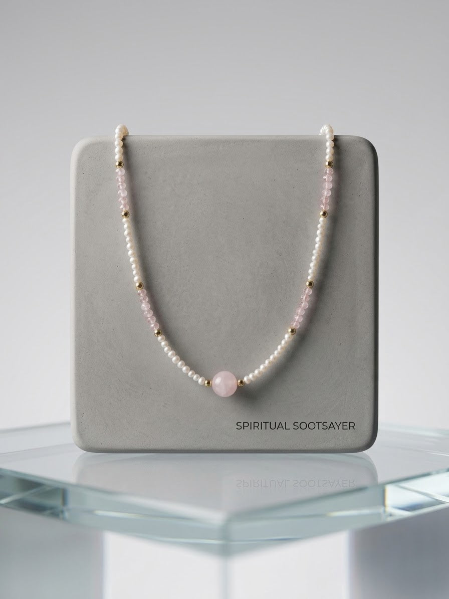 A beaded necklace with white pearls, light pink beads, and small gold spacer beads, featuring a central round light pink bead. The necklace is draped over a gray display card printed with the brand name 'SPIRITUAL SOOTSAYER', set on a clear glass display stand against a neutral gray background.