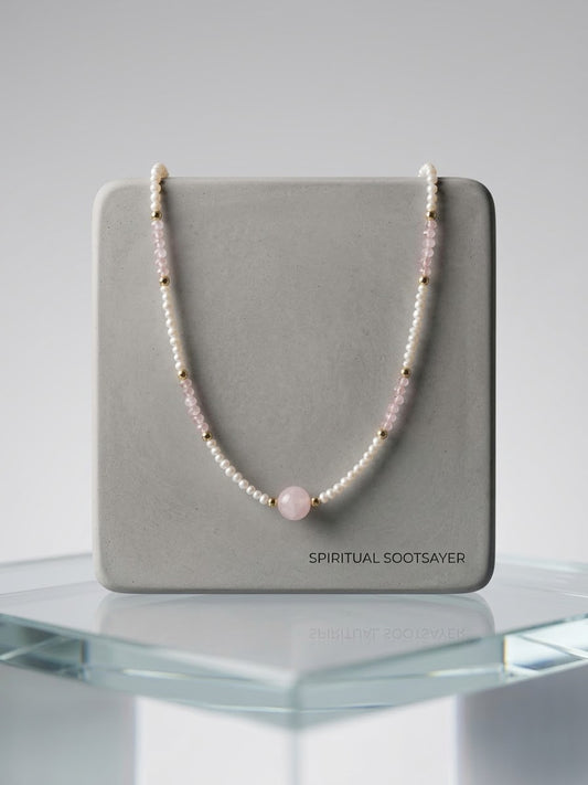A beaded necklace with white pearls, light pink beads, and small gold spacer beads, featuring a central round light pink bead. The necklace is draped over a gray display card printed with the brand name 'SPIRITUAL SOOTSAYER', set on a clear glass display stand against a neutral gray background.