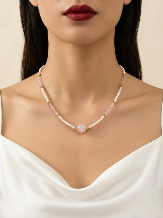 A close-up of a model wearing a delicate necklace made of small white pearls and pale pink beads, with a central round pink stone. The model is wearing a white satin top against a soft beige background.