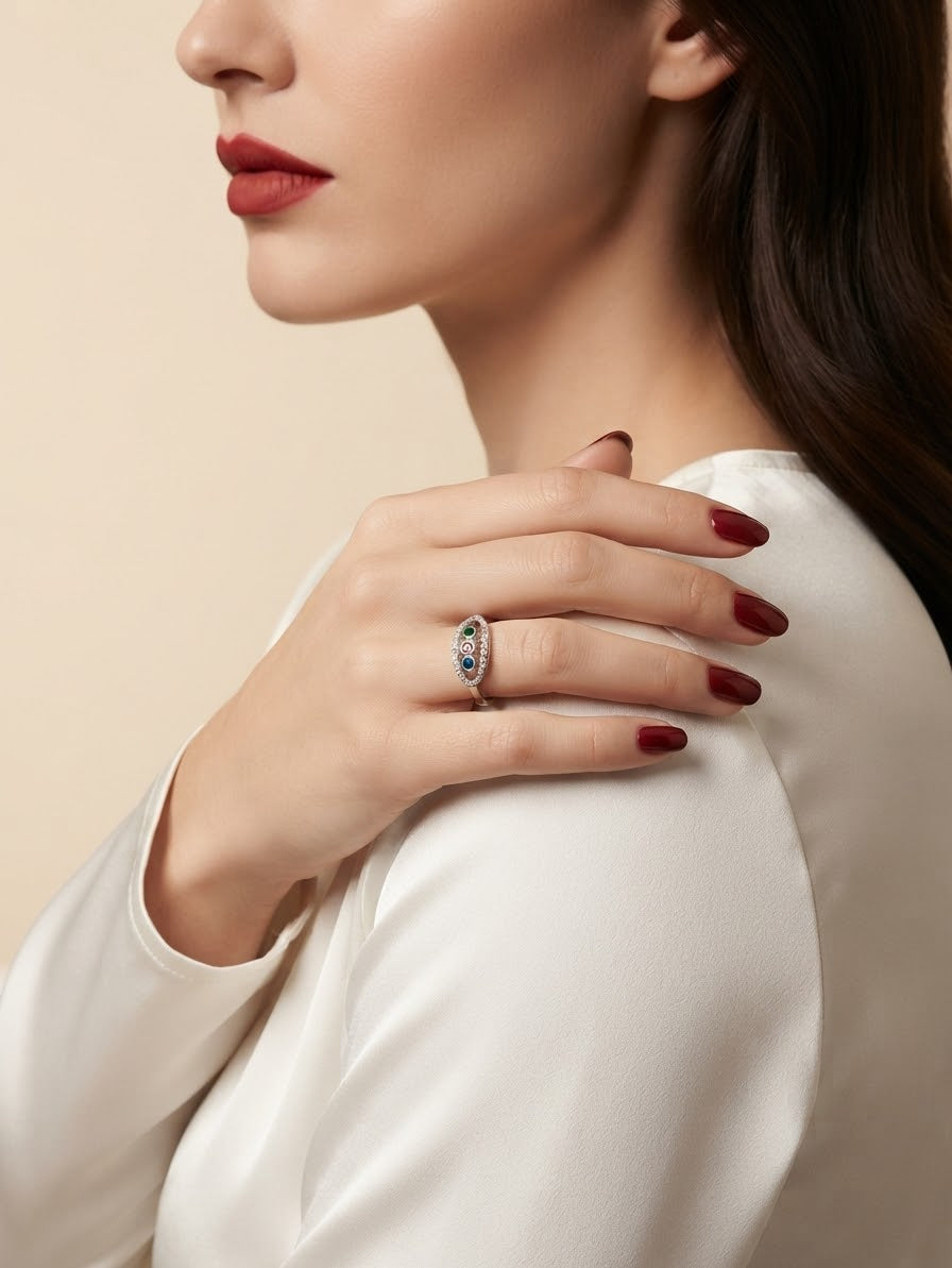 A close-up lifestyle shot of a woman wearing a silver ring with a blue and red stone cluster, styled with a white satin top and dark red nail polish against a warm beige background.