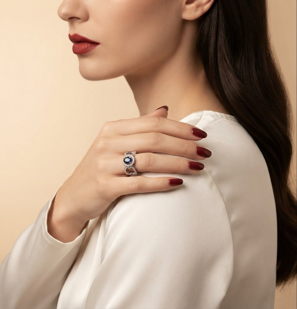 A close-up lifestyle shot of a model wearing a silver ring with a blue and white evil eye motif on her finger, paired with a cream satin top against a warm beige background.