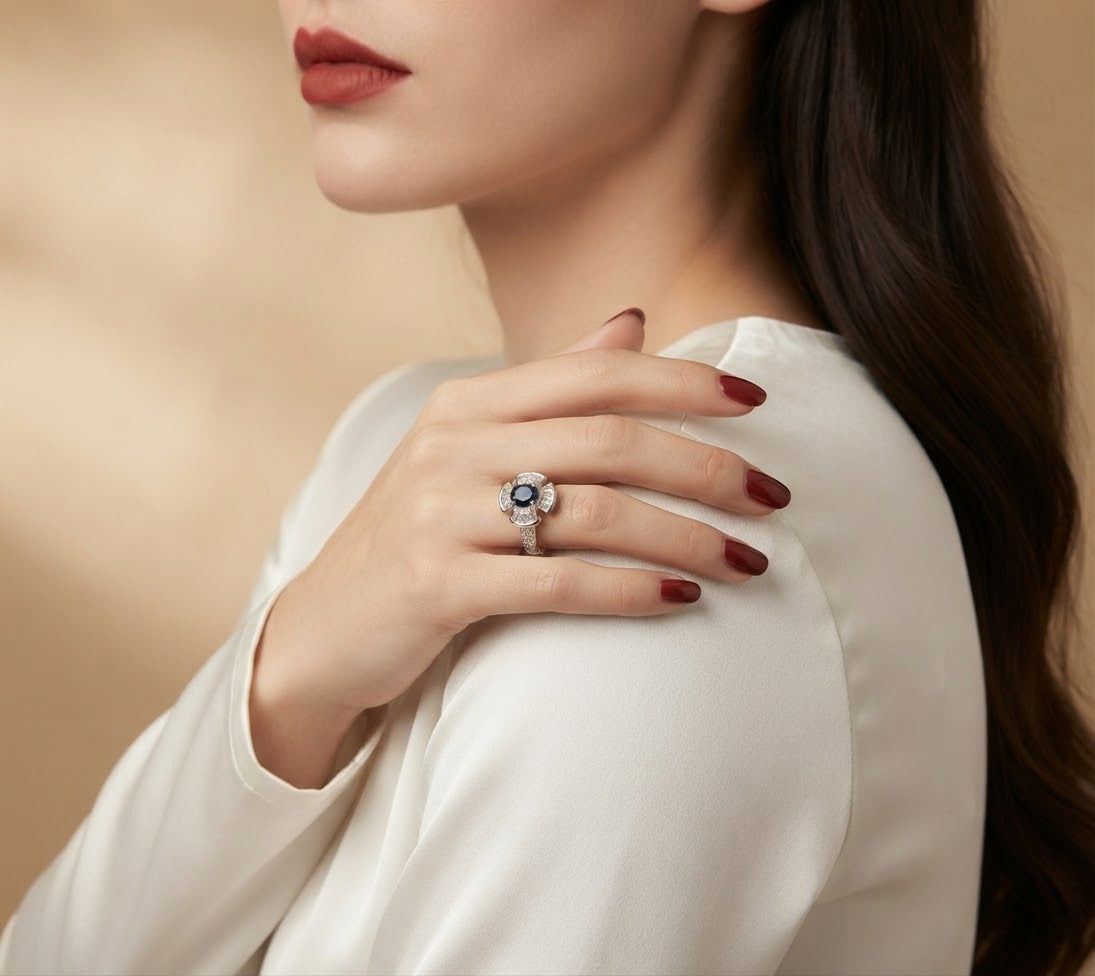 A close-up lifestyle shot of a woman wearing a silver-toned ring with a central blue gemstone and surrounding clear stones, styled with a white satin top against a warm beige background.