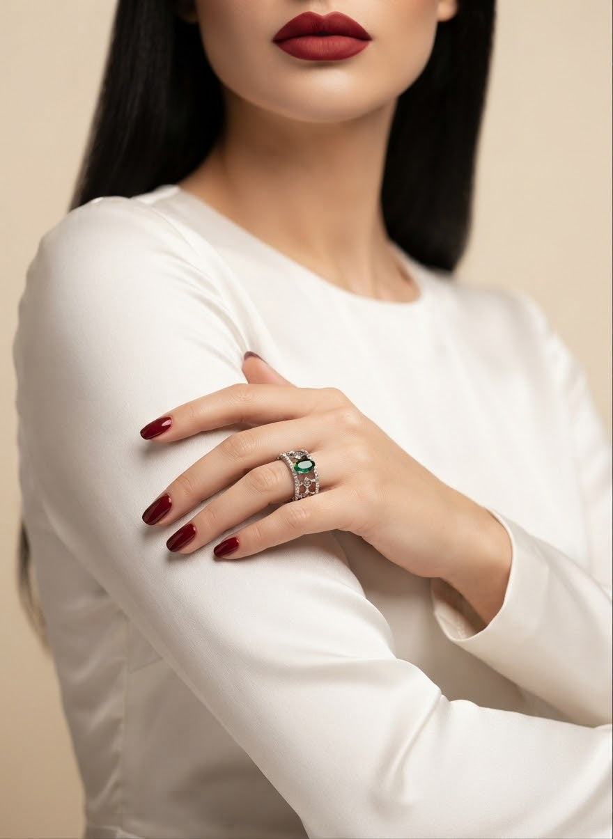 A close-up of a model wearing a silver ring with a green oval gemstone and small clear stones, paired with a white long-sleeve top and dark red lipstick.