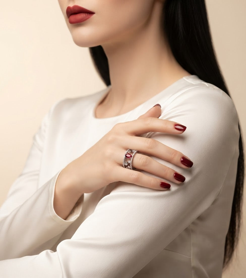 A close-up lifestyle shot of a woman wearing a silver ring with a red heart-shaped gemstone on her finger, dressed in a white long-sleeve top against a warm beige background.