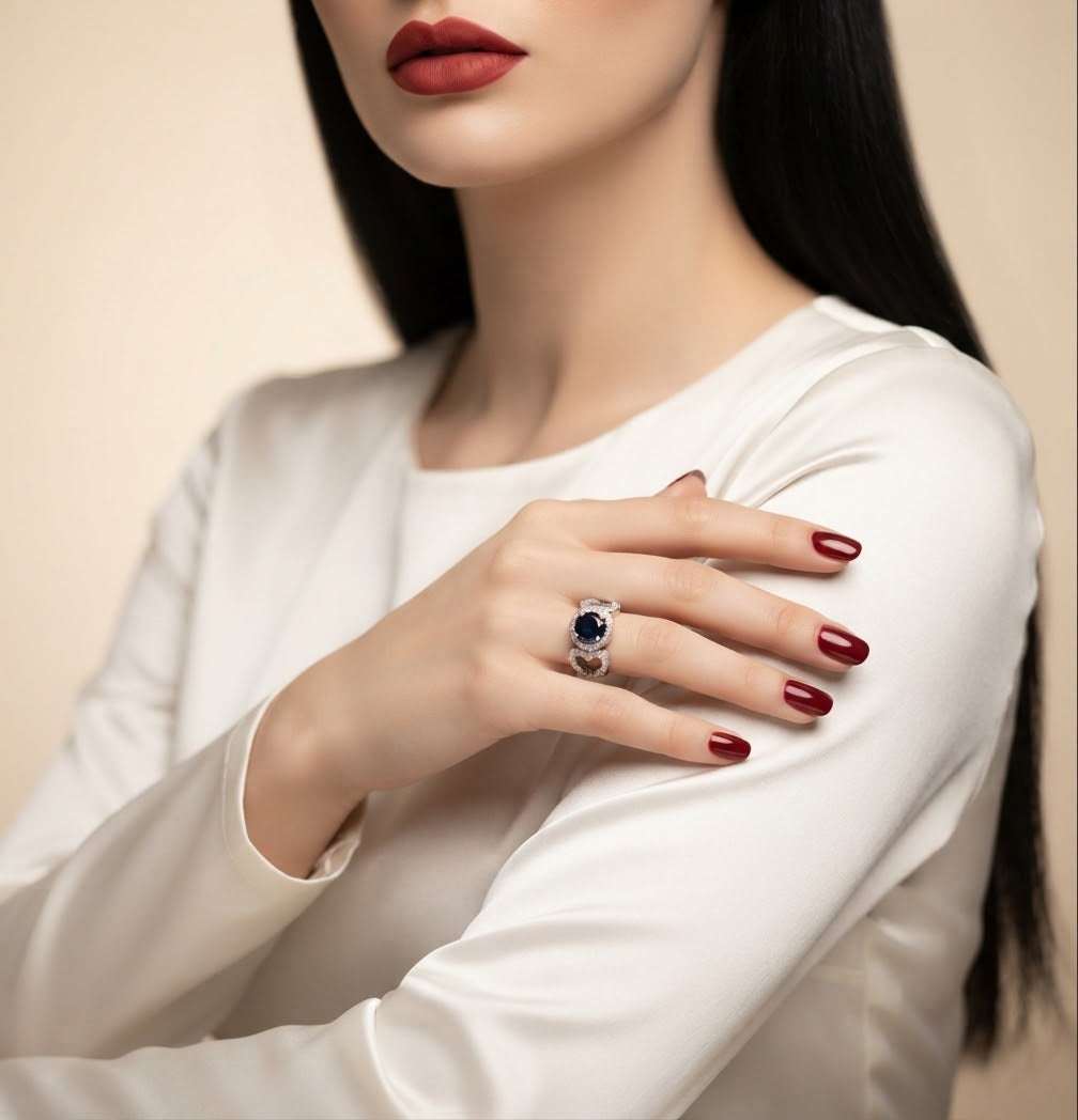 A close-up lifestyle shot of a model wearing a silver ring with a deep blue oval gemstone, styled with a white satin top and dark red nail polish against a soft beige background.