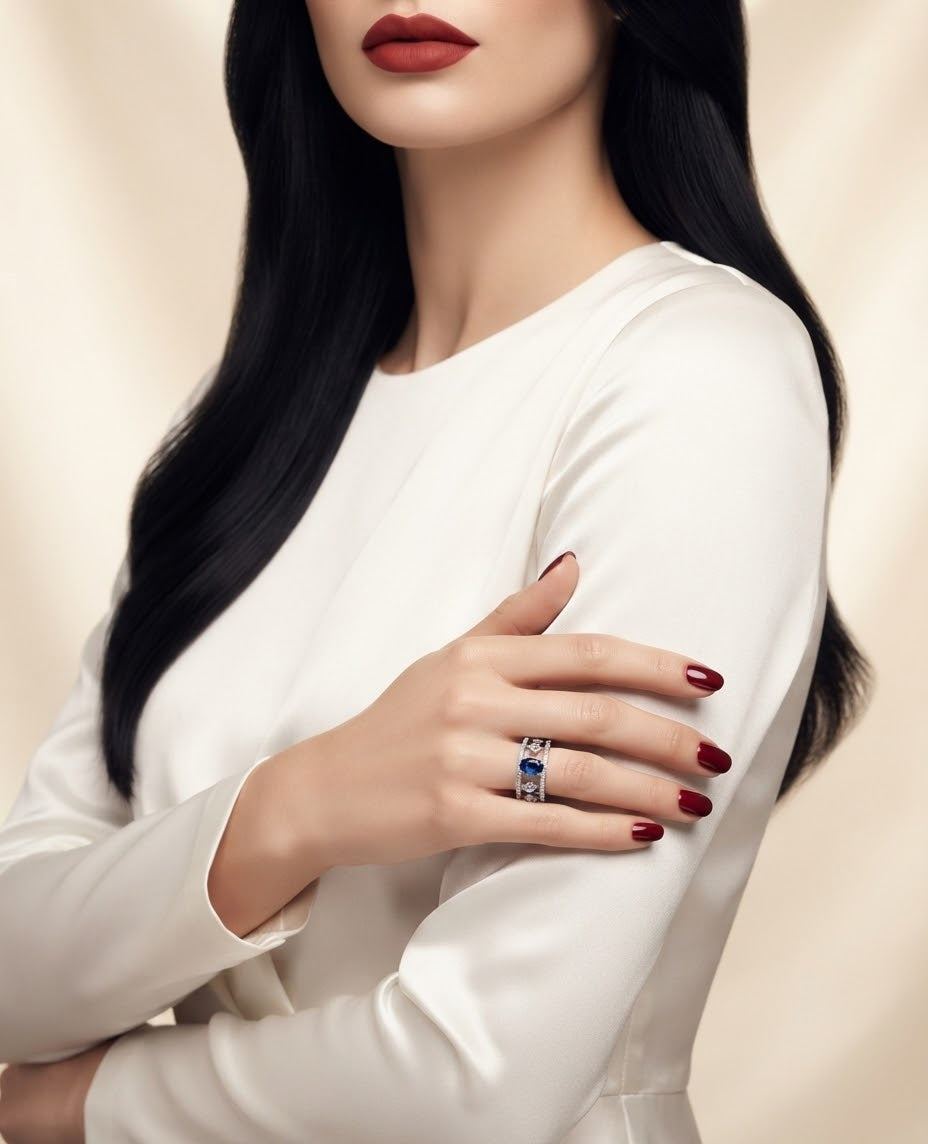 A close-up of a woman wearing a white long-sleeve top, showcasing a silver ring with blue gemstones on her finger. She has long black hair, red lipstick, and red manicured nails, posed against a soft beige background.