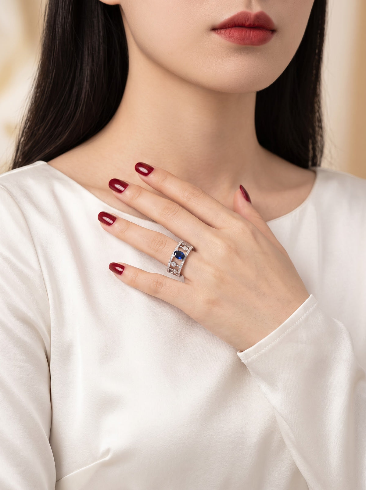 A close-up of a woman wearing a silver ring with a central deep blue oval gemstone, styled on her finger with dark red manicured nails and a white satin top.