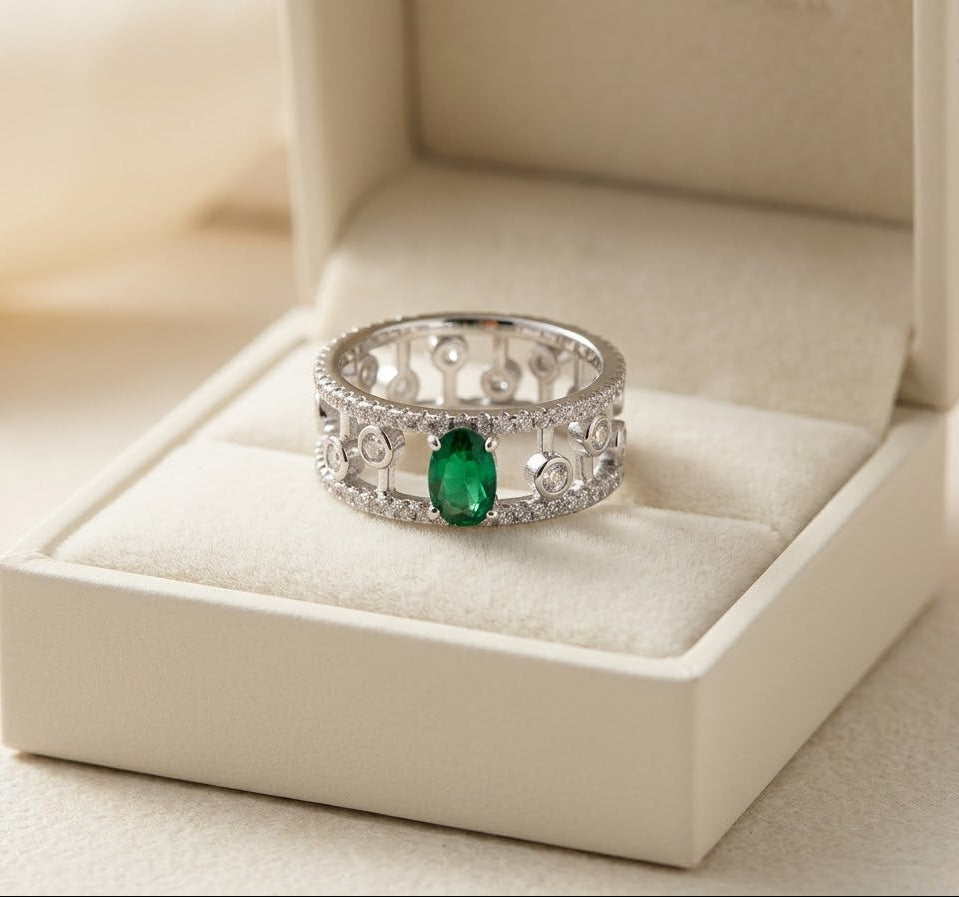 An ornate silver ring with a central green oval gemstone and small clear stones, displayed in an open cream-colored jewelry box.