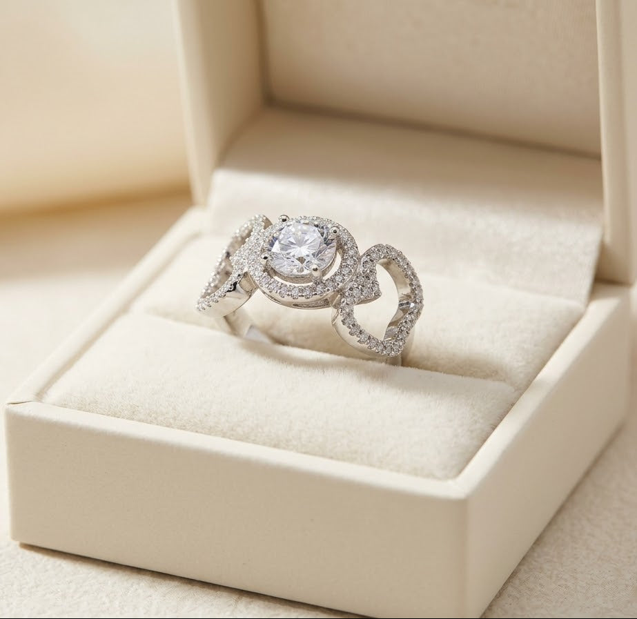 A silver ring with a large clear center stone and pavé-set curved side stones, displayed in an open cream-colored jewelry box.