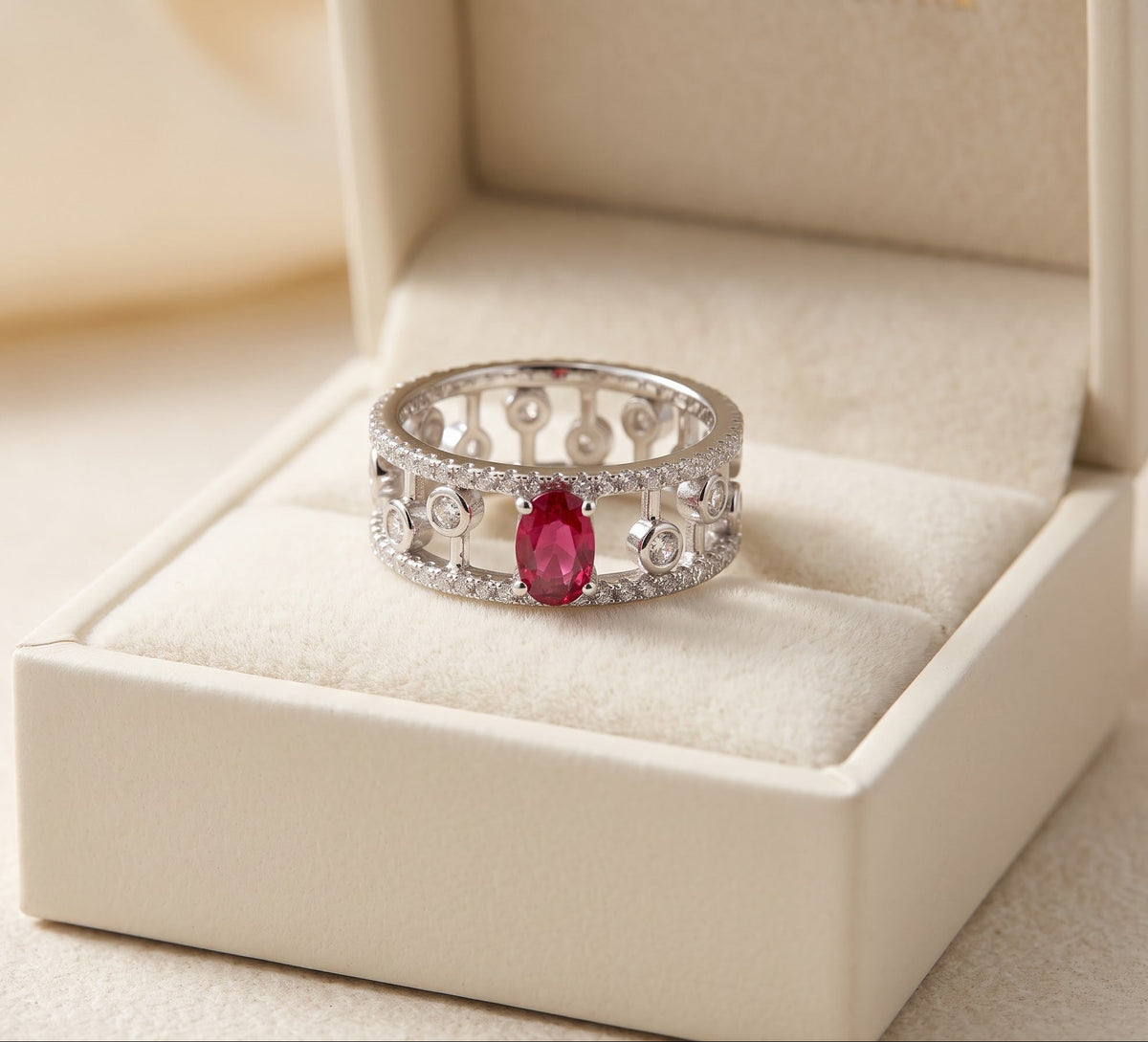 A silver filigree ring with a central marquise-cut red gemstone, displayed in an open cream-colored jewelry box.