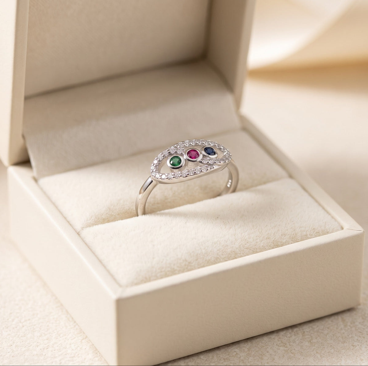 A silver ring with three small colored gemstones (green, red, and blue) surrounded by clear pavé stones, displayed in an open cream-colored jewelry box.