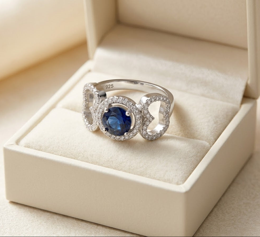 A silver ring with a central oval blue gemstone and diamond accents, displayed in an open cream-colored jewelry box.