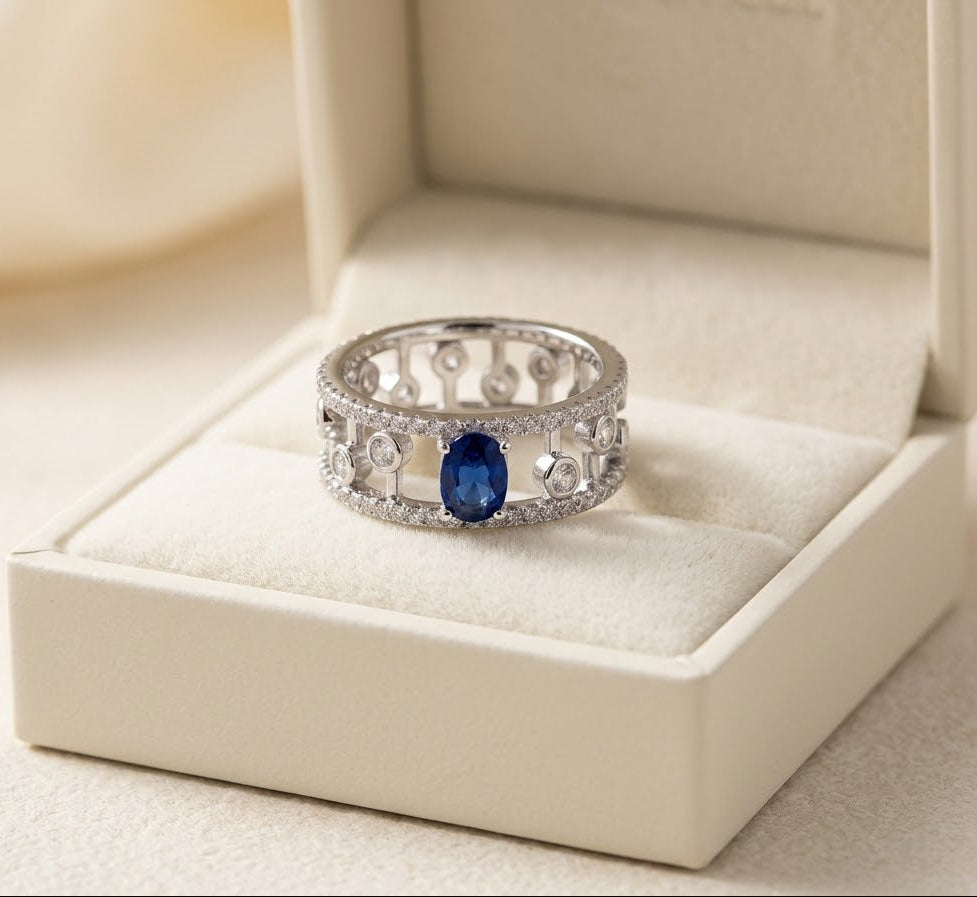 A silver filigree ring with a central oval blue gemstone and small clear stones, displayed in an open cream-colored jewelry box.