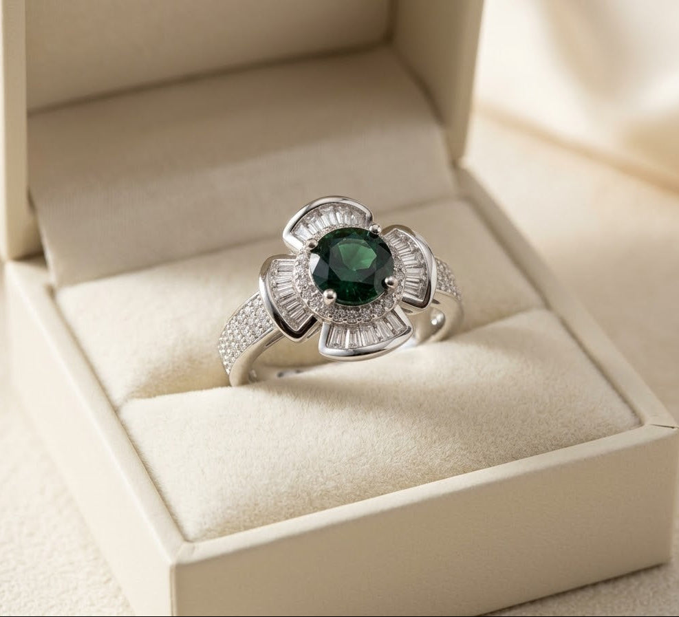 A silver-toned ring with a central round green gemstone and baguette-cut side stones, displayed in an open cream-colored jewelry box.