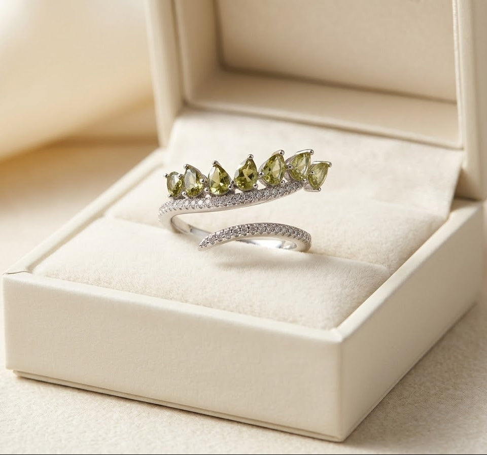 A silver ring with five marquise-cut olive green gemstones and a curved pavé band, displayed in an open cream-colored jewelry box.