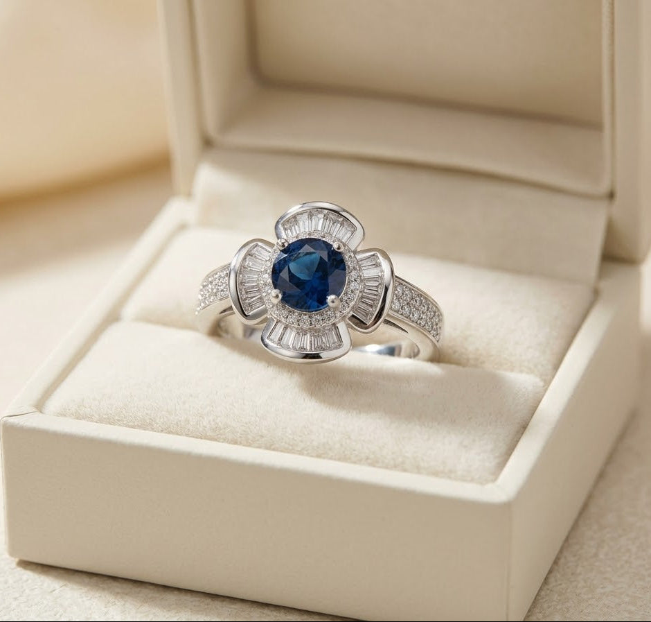 A silver ring with a central deep blue oval gemstone and surrounding clear stones, displayed in an open cream-colored jewelry box.