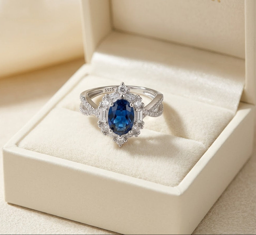 A silver ring with a central oval blue gemstone and small clear accent stones, displayed in an open cream-colored jewelry box.