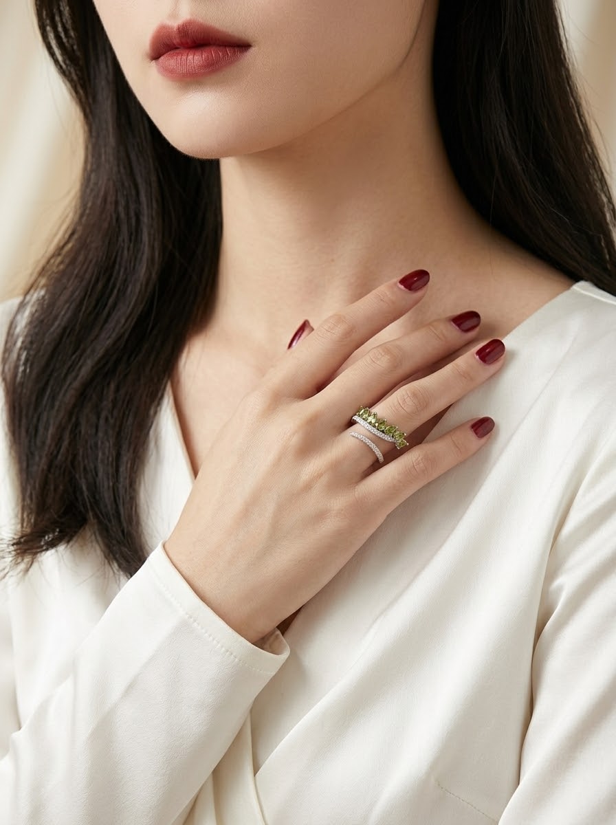 A close-up of a woman wearing a silver ring with a green stone cluster, styled on her finger with dark red nail polish and a white satin top.