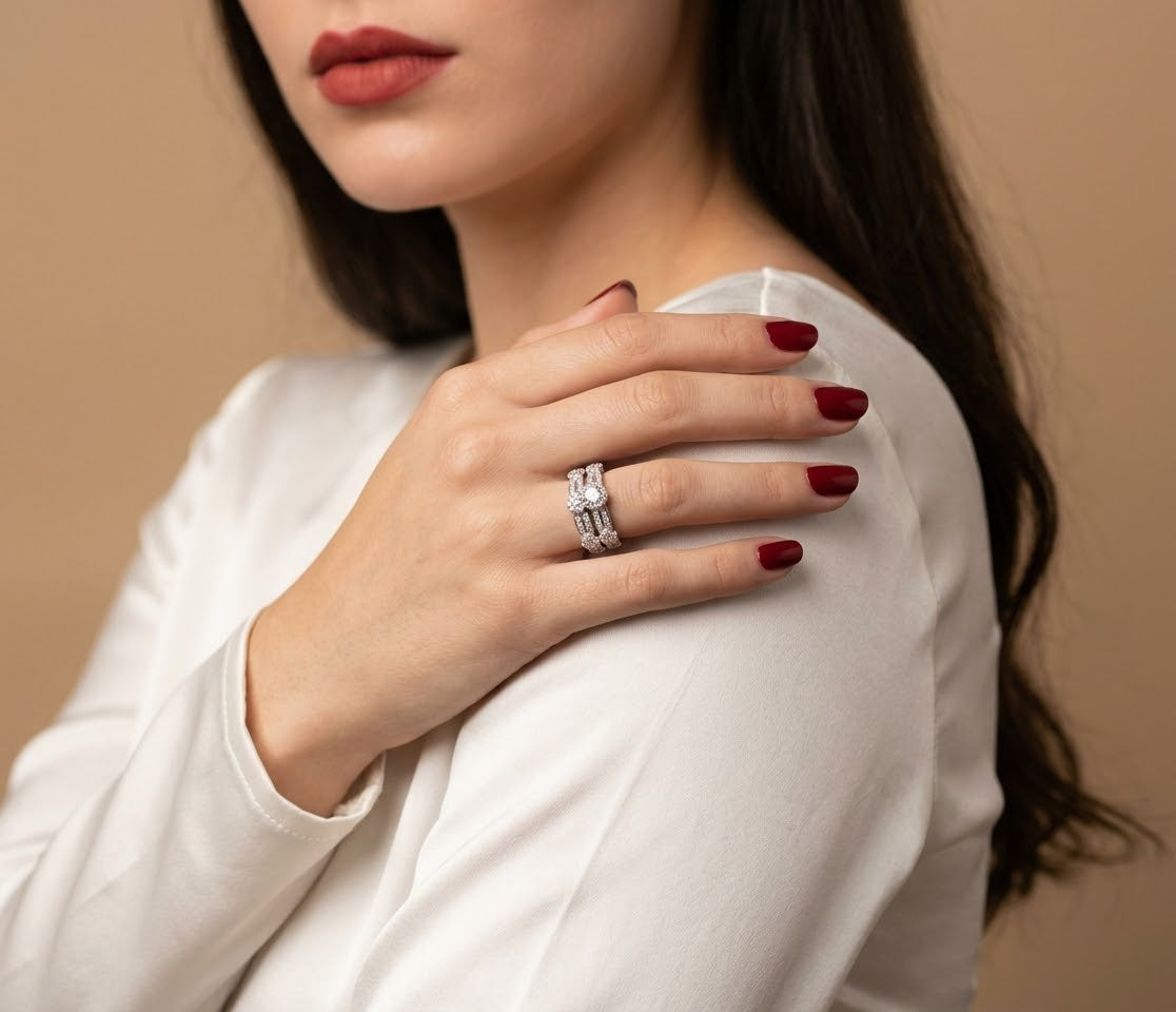 A close-up of a woman wearing a silver-toned double-band ring with clear stones, resting her hand on her shoulder. She is wearing a white long-sleeve top and has dark red nail polish against a solid beige background.