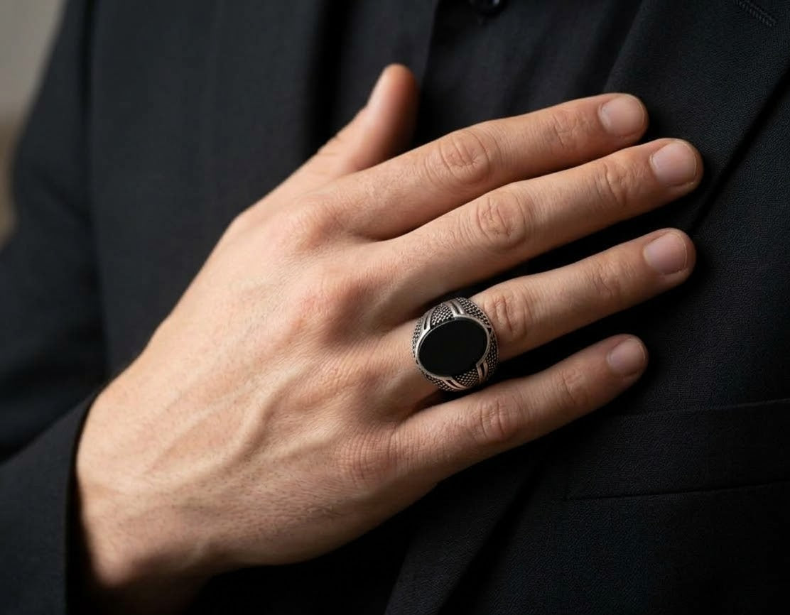 A close-up of a man's hand resting on his chest, wearing a silver signet ring with a large black oval face and textured band, set against a black suit jacket.