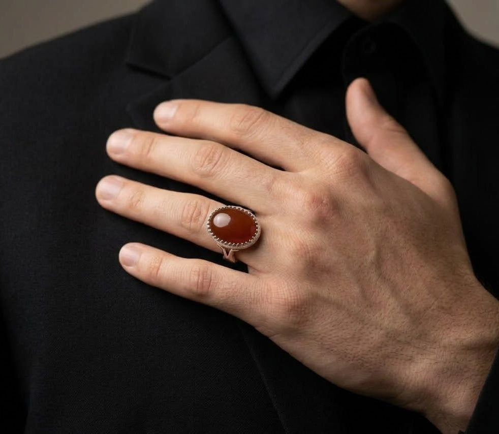 A close-up of a person's hand wearing a rose gold ring with a round brown cabochon stone, set against a black suit jacket and shirt.
