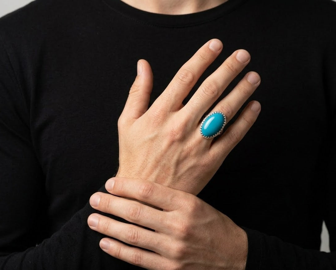A close-up of a person's hands wearing a silver ring with a vibrant turquoise oval stone, set against a black long-sleeve shirt.