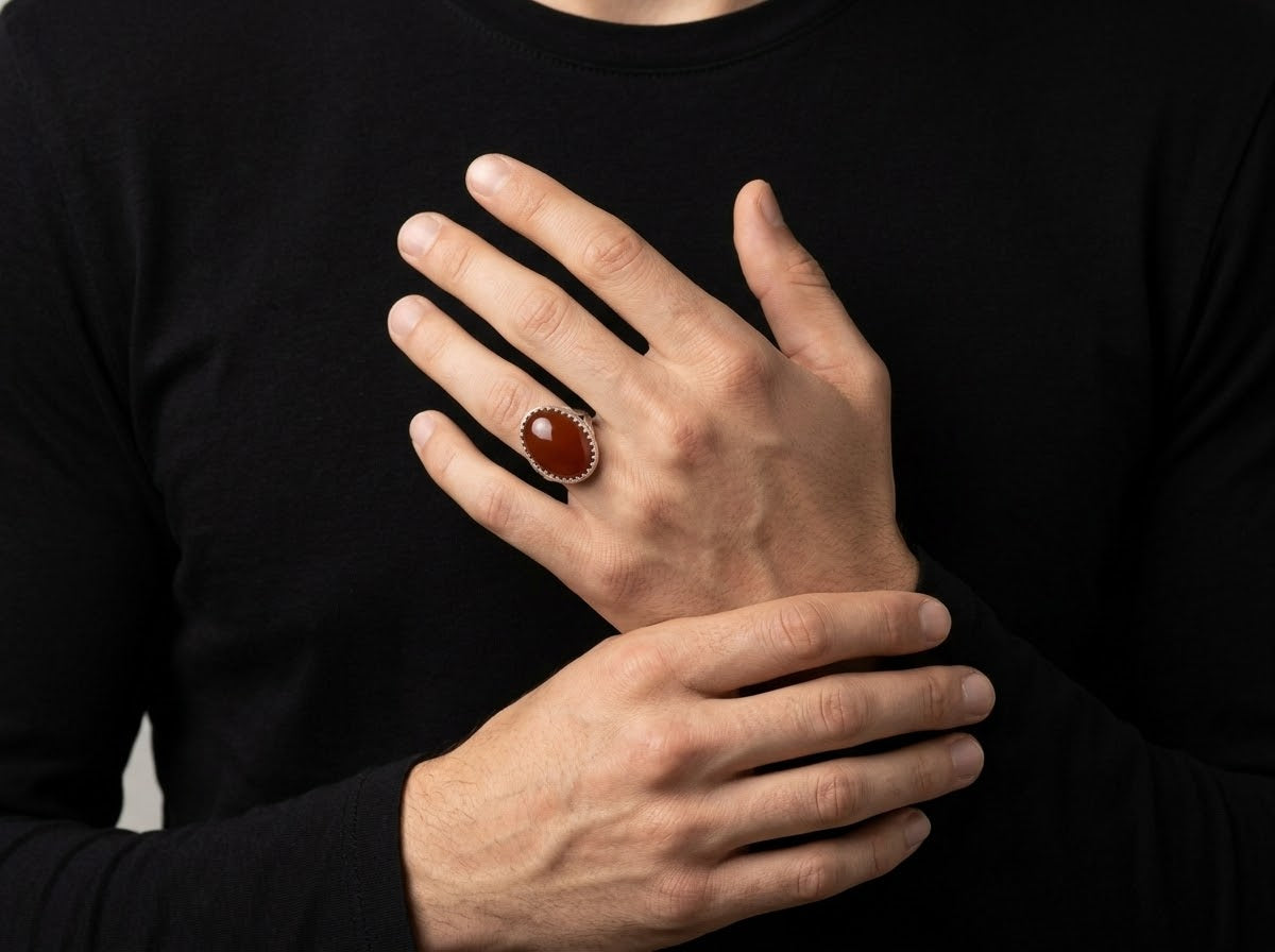 A close-up of a person wearing a silver-toned ring with a large oval brown cabochon stone, with their hands crossed over their chest against a black long-sleeve shirt.
