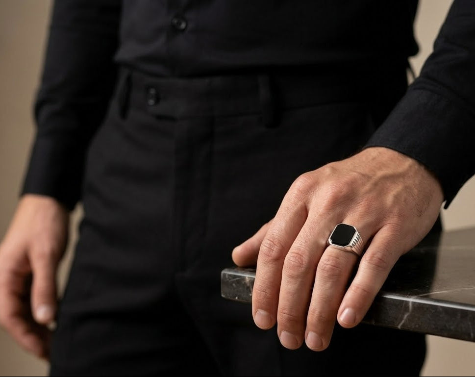 A close-up of a man's hand resting on a dark marble table, wearing a silver signet ring with a black rectangular face and bezel.