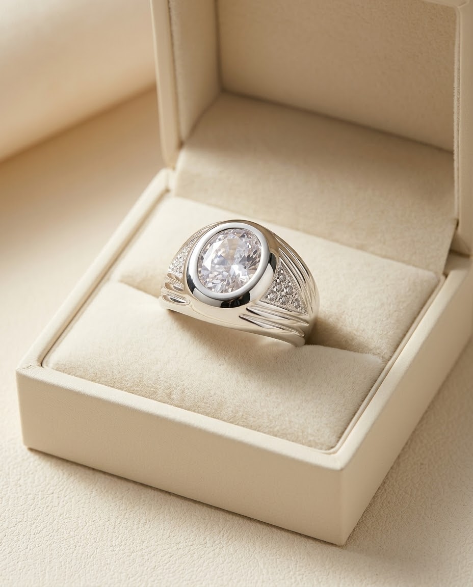 A silver signet-style ring with a large oval clear gemstone and small side stones, displayed in an open cream jewelry box.