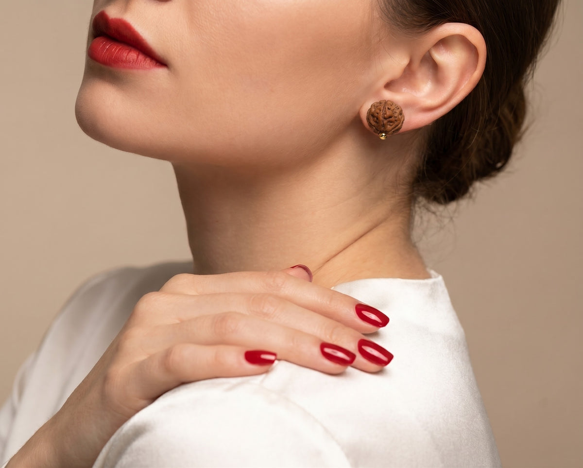 A close-up of a woman wearing a small round gold-toned stud earring with an intricate floral or scrollwork design, styled with red lipstick and red manicured nails.