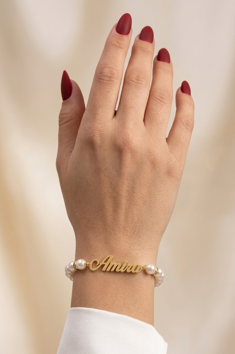 Hand wearing a gold bracelet with pearl accents and 'Amira' engraved on a beige background