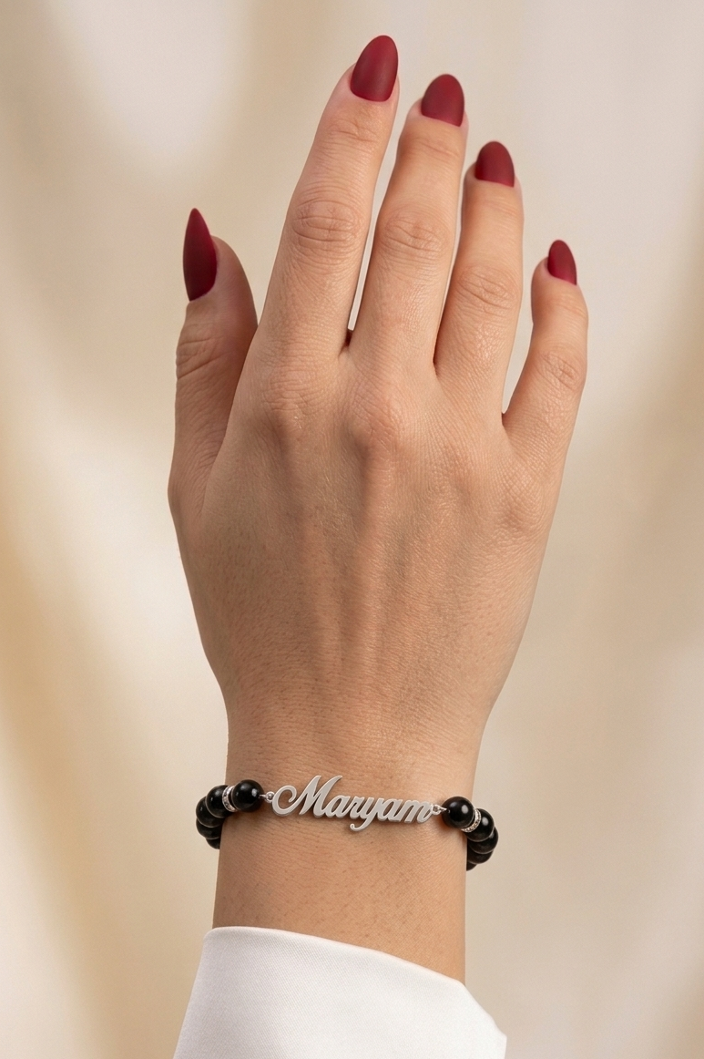 Hand wearing a black beaded bracelet with 'Maryam' engraved on a beige background