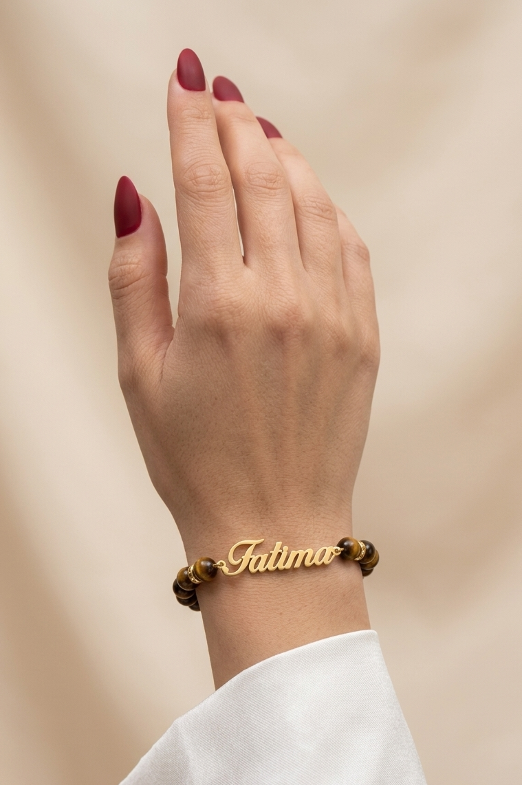 Hand wearing a gold bracelet with 'Fatima' engraved on it against a beige background