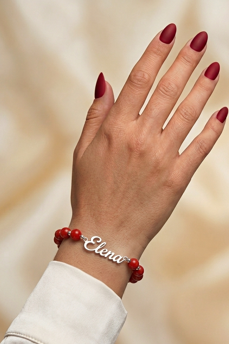 Hand wearing a bracelet with red beads and 'Elena' engraved on a beige background