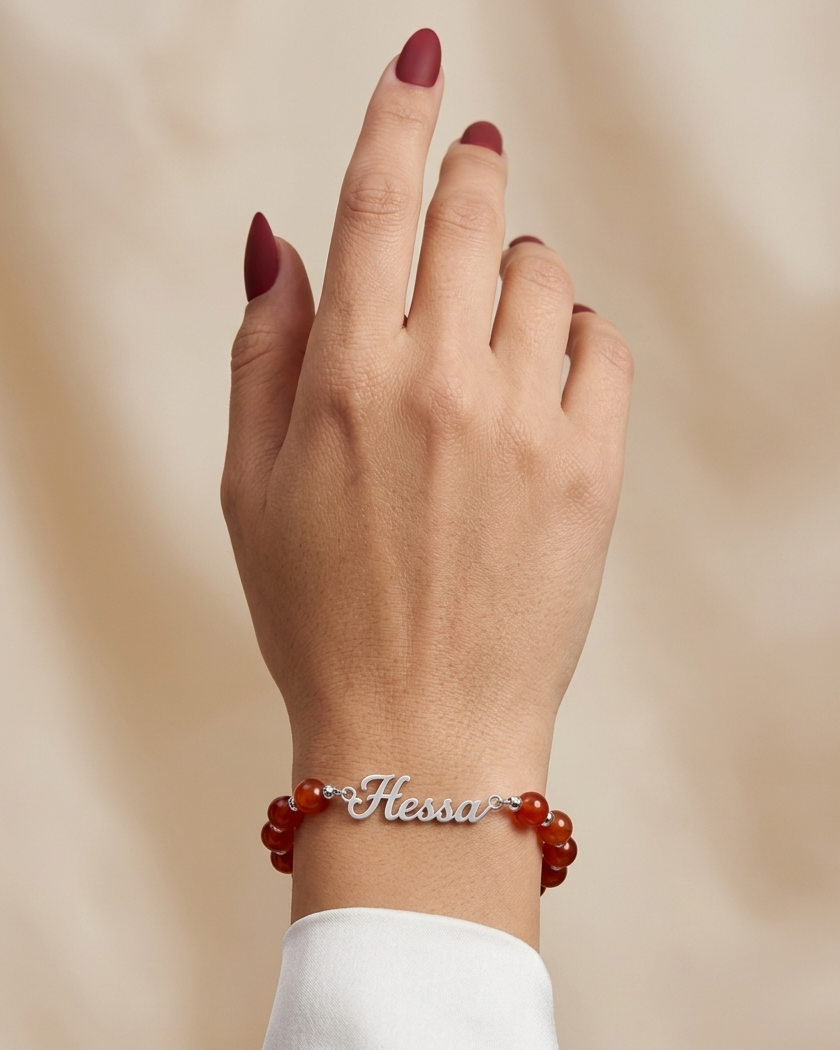 Hand wearing a bracelet with red beads and 'Hessa' engraved on a beige background