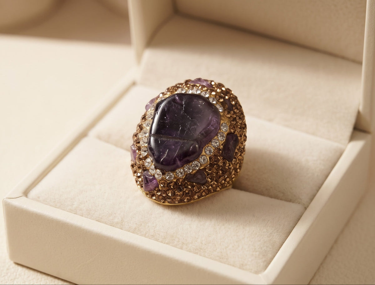 A gold ring with a large oval purple amethyst cabochon and smaller clear stones, displayed in an open cream-colored jewelry box.
