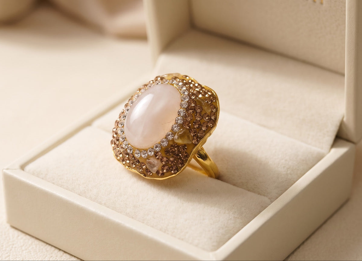 A gold ring with a large oval pale pink stone and small clear stones around it, displayed inside an open cream-colored jewelry box.