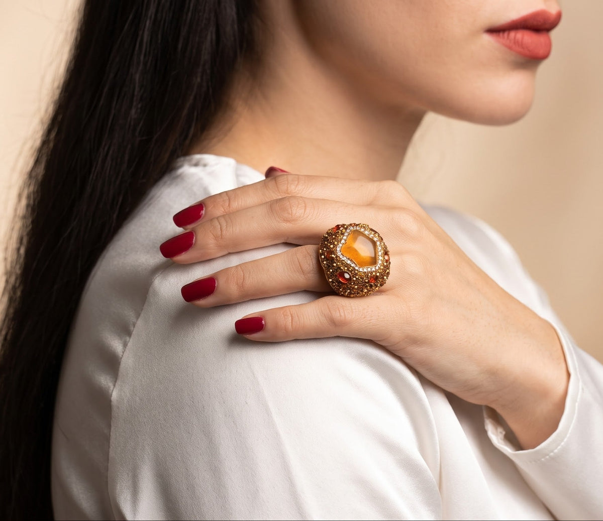 A close-up lifestyle shot of a woman wearing a large statement ring on her hand, which is resting on her shoulder. The ring features a prominent oval orange stone surrounded by a textured gold setting.