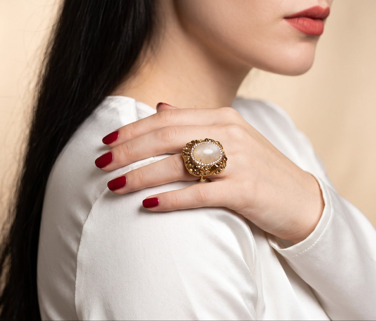 A close-up lifestyle shot of a woman wearing a large gold ring with a round white stone, resting her hand on her shoulder.