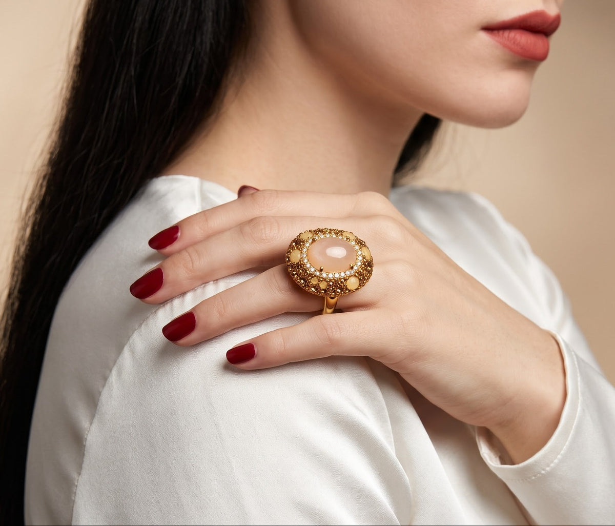A close-up of a woman wearing a large gold ring with a pale pink stone on her hand, which is resting on her shoulder. She is wearing a white top and has dark red manicured nails, set against a soft beige background.
